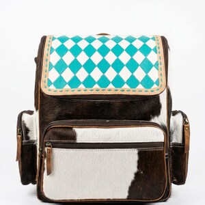 May include: A cowhide backpack with a turquoise and white diamond pattern on the flap. The bag features brown leather trim, a front zippered pocket, and side pockets. The main body of the backpack is brown and white cowhide.