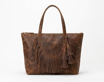May include: A brown leather tote bag featuring a semi-circular fringe panel on the front, two shoulder straps, and a decorative tassel charm attached to one side. The bag has a top zipper closure and a smooth, matte finish with natural variations.