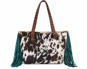 May include: A tote bag featuring a black and white cowhide pattern on the front and back. The sides are adorned with long, teal-colored fringe. It includes two brown leather shoulder straps decorated with small, evenly spaced silver metal studs.