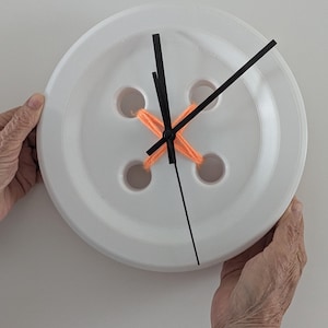 May include: A white, button-shaped wall clock with black hands and orange yarn detail. The clock has four holes and is held by two hands against a white background. A unique, decorative timepiece.