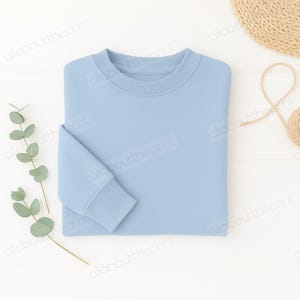 May include: A pale blue crew neck sweatshirt, folded. The sleeves have ribbed cuffs, and the collar is ribbed. A sprig of eucalyptus and a woven coaster are in the background.