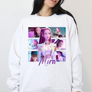 May include: A light gray sweatshirt featuring a graphic of a woman with purple hair and the name "Mira" in cursive. The graphic is a collage of images. The sweatshirt is a casual style.