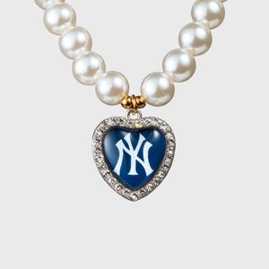 Könnte beinhalten: Eine Perlenkette mit herzförmigem Anhänger. Der Anhänger hat einen blauen Hintergrund mit weißem "NY"-Logo, umgeben von einem silberfarbenen Rand. Die Kette besteht aus großen, runden, weißen Perlen.
