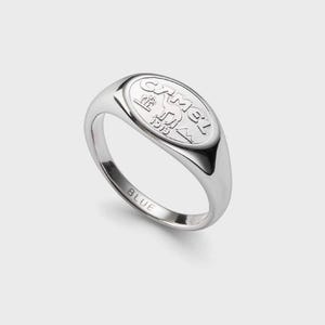 Puede incluir: Un anillo de sello plateado con una cara ovalada. La cara presenta la palabra "Camel" sobre un camello y un diseño de pirámide, con el año "1913" debajo. La palabra "BLUE" está grabada en la banda.