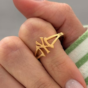 May include: A gold ring with a distinctive design. The ring is held by a hand with long, polished nails. The ring's design is a series of connected shapes. The hand is wearing a green and white striped sleeve.