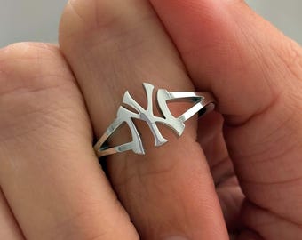 May include: A silver ring featuring the stylized letters "NY" in a cut-out design. The ring is shown on a finger with a light pink manicure. The ring's design is open, with the letters connected by thin bands of metal.