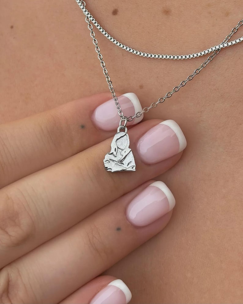 K&ouml;nnte beinhalten: Eine silberne Halskette mit zwei Ketten und einem Anh&auml;nger. Der Anh&auml;nger ist eine kleine, detaillierte Figur einer Person. Die Halskette wird am Hals einer Person getragen, mit der Hand und den N&auml;geln im Bild. Die N&auml;gel sind mit French Manicure lackiert.