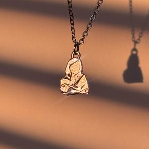 Peut inclure: Un collier avec un pendentif dor&eacute; repr&eacute;sentant une silhouette humaine. Le pendentif est suspendu &agrave; une cha&icirc;ne sombre. L'arri&egrave;re-plan est de couleur neutre et chaude avec des ombres.