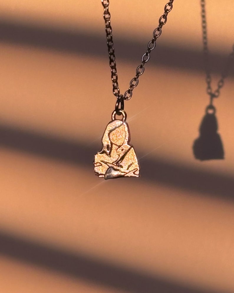 Peut inclure: Un collier avec un pendentif dor&eacute; repr&eacute;sentant une silhouette humaine. Le pendentif est suspendu &agrave; une cha&icirc;ne sombre. L'arri&egrave;re-plan est de couleur neutre et chaude avec des ombres.