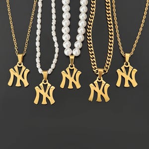 Op de afbeelding: Een verzameling goudkleurige kettingen met hangers met de letters "NY". De kettingen hebben verschillende kettingstijlen, waaronder een klassieke ketting, een parelketting en een gourmetteketting. De achtergrond is effen zwart.