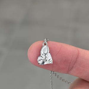K&ouml;nnte beinhalten: Eine kleine, silberfarbene Anh&auml;ngerkette mit einem stilisierten Bild einer Frau. Der Anh&auml;nger ist an einer filigranen Kette befestigt. Die Halskette wird zwischen zwei Fingern vor einem verschwommenen grauen Hintergrund gehalten.