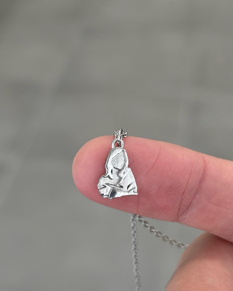 K&ouml;nnte beinhalten: Eine kleine, silberfarbene Anh&auml;ngerkette mit einem stilisierten Bild einer Frau. Der Anh&auml;nger ist an einer filigranen Kette befestigt. Die Halskette wird zwischen zwei Fingern vor einem verschwommenen grauen Hintergrund gehalten.