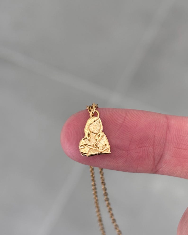 Peut inclure: Un collier pendentif dor&eacute; avec une silhouette stylis&eacute;e d'une personne. Le pendentif est une petite figurine d&eacute;taill&eacute;e avec les bras crois&eacute;s. La cha&icirc;ne du collier est d&eacute;licate et dor&eacute;e, avec un fermoir simple.