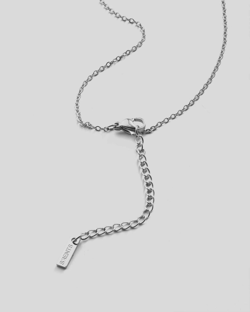 K&ouml;nnte beinhalten: Eine silberfarbene Halskette mit einer filigranen Kette und einem Karabinerverschluss. Die Halskette hat eine Verl&auml;ngerungskette mit einem kleinen rechteckigen Anh&auml;nger mit der Aufschrift "S.HENTS". Der Schmuck wird vor einem hellgrauen Hintergrund pr&auml;sentiert.