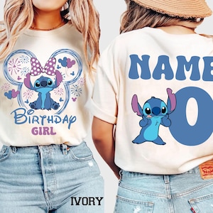 May include: Ivory-colored t-shirt featuring a blue cartoon character with a bow, surrounded by a Mickey Mouse-shaped design with fireworks and the text "Birthday Girl." The back of the shirt has the word "NAME" and the number "0".