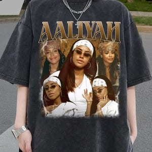 May include: Dark grey oversized t-shirt with a graphic of Aaliyah. The graphic features multiple images of Aaliyah and the name "AALIYAH" in a gold font. The shirt has a vintage, washed appearance.