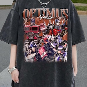 Könnte beinhalten: Dunkelgraues T-Shirt mit einem Aufdruck von Optimus Prime und anderen Transformers-Charakteren. Der Text "OPTIMUS Prime" ist in Braun und Orange gehalten. Das T-Shirt hat einen Vintage-Look.