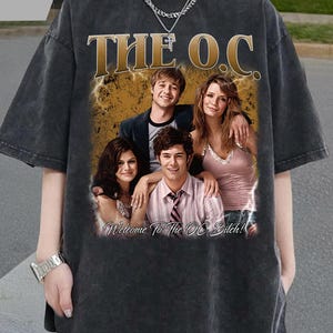 Puede incluir: Camiseta gris oscuro con un gráfico vintage del elenco de "The O.C.". El diseño incluye el título del programa en letras doradas y la frase "Welcome to The O.C. Bitch!" debajo de la foto del elenco.