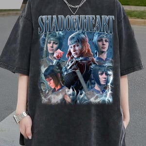 Puede incluir: Camiseta gris oscuro de gran tamaño con la palabra "SHADOWHEART" en texto azul y blanco. La camiseta muestra un gráfico de varios personajes de fantasía. Tiene mangas cortas y cuello redondo.