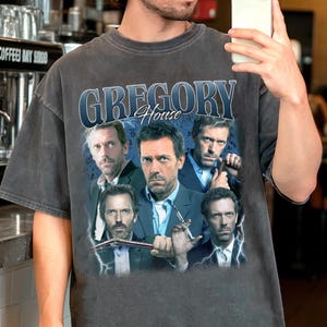 以下が含まれることがあります： テレビドラマ「House」のグラフィックが特徴の濃いグレーのTシャツ。「Gregory House」という番組タイトルとキャストの画像が含まれています。シャツはヴィンテージスタイルで、少し色あせた外観です。
