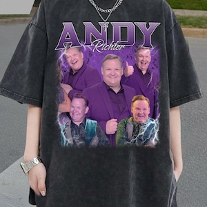 Puede incluir: Camiseta gris oscuro con un gráfico de Andy Richter en varias poses, y el nombre "ANDY Richter" en texto morado. La camiseta tiene un aspecto vintage y descolorido.