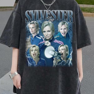 May include: Dark gray t-shirt featuring a graphic of the character Sue Sylvester from the TV show Glee. The graphic includes multiple images of the character and the text "SYLVESTER Sue". The shirt has a vintage, distressed look.