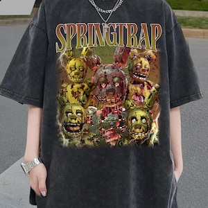 May include: Dark gray t-shirt featuring a graphic of the character Springtrap from the video game series Five Nights at Freddy's. The word "SPRINGTRAP" is printed across the top of the graphic in yellow and green.