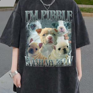 Pode incluir: T-shirt cinzento escuro oversized com um gráfico de cachorros e o texto "I'M PIBBLE WASH MY BELLY". A camisa tem um aspeto vintage e lavado, com mangas curtas. A marca "TAAJIRBILLAH" está impressa no canto superior esquerdo.