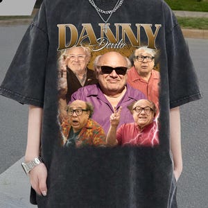 May include: Dark gray t-shirt featuring a collage of images of Danny DeVito. The shirt has the name "DANNY DeVito" in gold lettering. The shirt is a vintage style, with a slightly faded appearance.