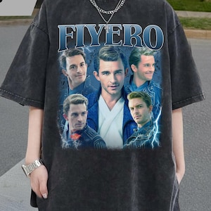 May include: Dark gray t-shirt featuring the word "FIYERO" in blue, with a collage of male portraits. The shirt has a vintage, washed-out look. The text "TAAJIRBILLAH" is in the upper left corner.
