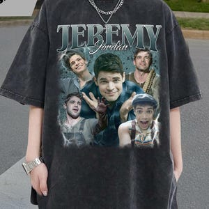 以下が含まれることがあります： ダークグレーのオーバーサイズTシャツ。コラージュ画像と「JEREMY Jordan」の文字がスタイリッシュなフォントでプリントされています。シャツはヴィンテージ感のある色褪せた外観です。柔らかく快適な素材で作られています。