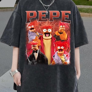 Könnte beinhalten: Dunkelgraues T-Shirt mit einer Grafik der Muppet-Figur Pepe the King Prawn. Die Grafik enthält das Wort "PEPE" in roten Buchstaben und mehrere Bilder der Figur. Das Shirt hat kurze Ärmel und einen Rundhalsausschnitt.