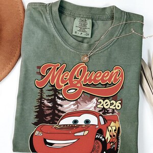 Puede incluir: Una camiseta de cuello redondo color verde oliva con un estampado gr&aacute;fico de un coche de carreras rojo con el n&uacute;mero 95, pinos y monta&ntilde;as de fondo. El texto "McQueen" aparece en una fuente roja grande sobre el coche, con "2026" a la derecha.