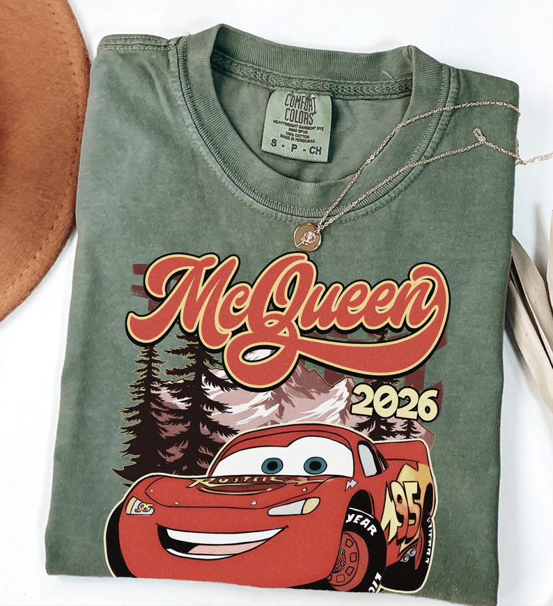 Puede incluir: Una camiseta de cuello redondo color verde oliva con un estampado gr&aacute;fico de un coche de carreras rojo con el n&uacute;mero 95, pinos y monta&ntilde;as de fondo. El texto "McQueen" aparece en una fuente roja grande sobre el coche, con "2026" a la derecha.
