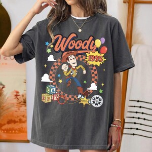 May include: A charcoal gray short-sleeve t-shirt featuring a graphic of the character Woody from Toy Story. The design includes the text Woody and 1995, colorful balloons, alphabet blocks, a sheriff star badge, and a film reel icon.