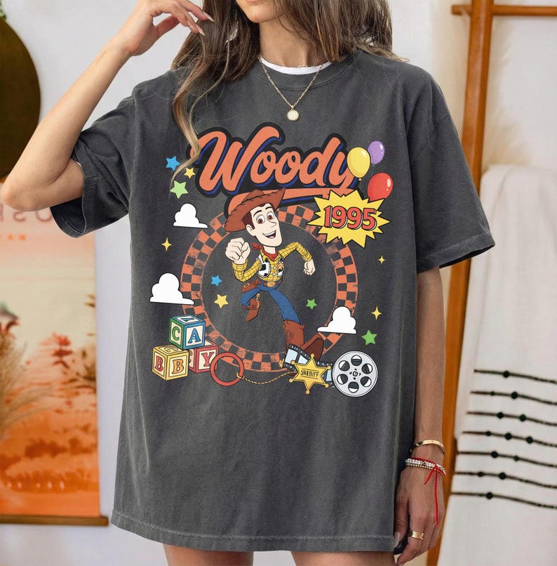 May include: A charcoal gray short-sleeve t-shirt featuring a graphic of the character Woody from Toy Story. The design includes the text Woody and 1995, colorful balloons, alphabet blocks, a sheriff star badge, and a film reel icon.