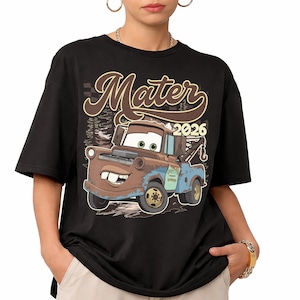 May include: A black short-sleeve t-shirt featuring a graphic of the character Mater from the movie Cars. The design includes the text "Mater" in a brown script font above the character and the year "2026" in a bold, light-colored font.