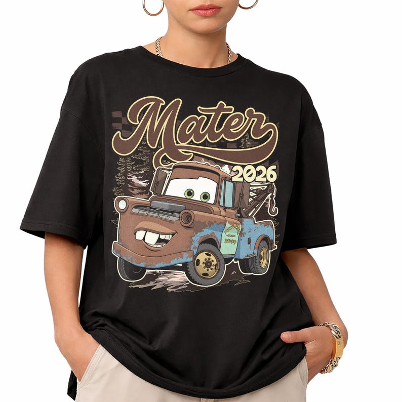 May include: A black short-sleeve t-shirt featuring a graphic of the character Mater from the movie Cars. The design includes the text "Mater" in a brown script font above the character and the year "2026" in a bold, light-colored font.