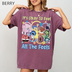 以下が含まれることがあります： ベリー色の半袖Tシャツで、様々なアニメキャラクターのグラフィックプリントが施されています。キャラクターの上には「It's Okay To Feel」、下には「All The Feels」という文字がプリントされており、カラフルな花のアクセントで囲まれています。