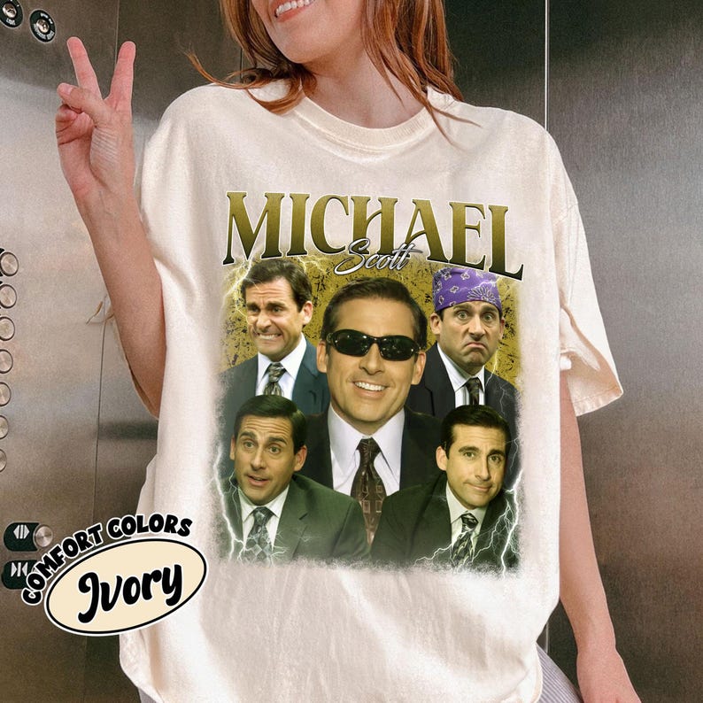 May include: Ivory-colored t-shirt featuring a collage of Michael Scott from The Office. The design includes the name "Michael Scott" in gold lettering above multiple images of the character. The shirt also has the words "Comfort Colors" and "Ivory" printed on it.