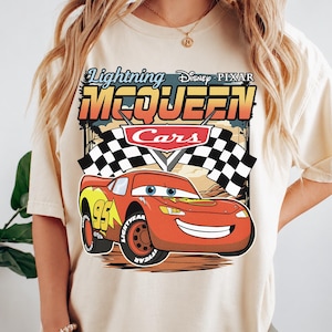 May include: A cream-colored t-shirt featuring a graphic of Lightning McQueen from Disney Pixar Cars. The design includes the text Lightning McQueen, the Cars logo, a checkered flag, and the red race car with the number 95 on its side.