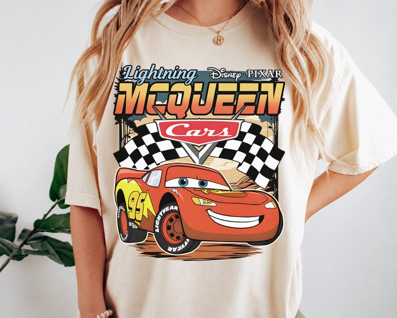 May include: A cream-colored t-shirt featuring a graphic of Lightning McQueen from Disney Pixar Cars. The design includes the text Lightning McQueen, the Cars logo, a checkered flag, and the red race car with the number 95 on its side.
