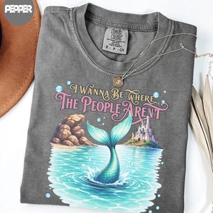 K&ouml;nnte beinhalten: Ein anthrazitfarbenes T-Shirt mit einem Grafikdruck, der eine Meerjungfrauenflosse zeigt, die aus blauem Wasser ragt, sowie eine felsige K&uuml;ste und ein Schloss. Der Text lautet I WANNA BE WHERE THE PEOPLE ARENT in rosa und goldener Schrift.