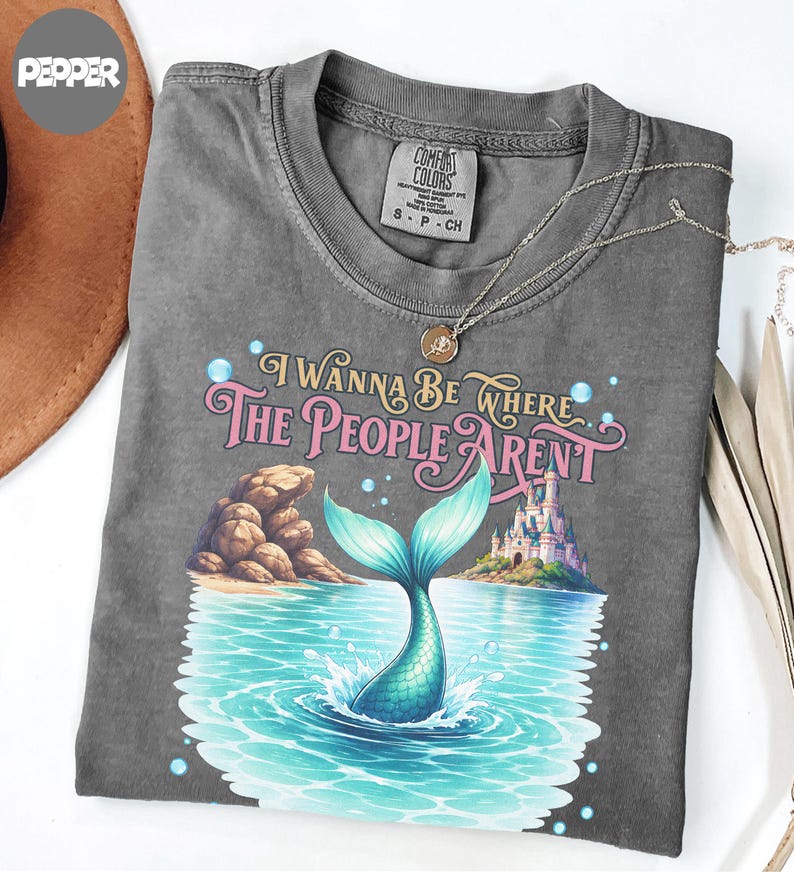 K&ouml;nnte beinhalten: Ein anthrazitfarbenes T-Shirt mit einem Grafikdruck, der eine Meerjungfrauenflosse zeigt, die aus blauem Wasser ragt, sowie eine felsige K&uuml;ste und ein Schloss. Der Text lautet I WANNA BE WHERE THE PEOPLE ARENT in rosa und goldener Schrift.