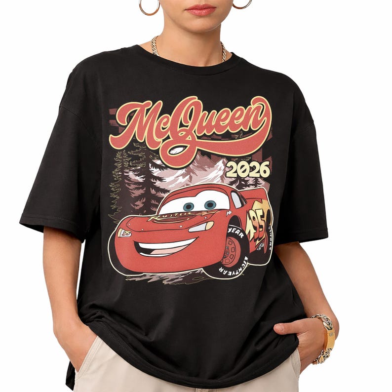 Puede incluir: Una camiseta negra de manga corta con un estampado gr&aacute;fico de un coche de carreras rojo con el texto McQueen y 2026. El dise&ntilde;o incluye un fondo de bosque estilizado detr&aacute;s del coche, con el n&uacute;mero 95 visible en el lateral del veh&iacute;culo.
