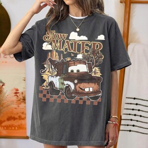 May include: A charcoal gray short-sleeve t-shirt featuring a graphic print of the character Tow Mater. The design includes the text "Tow Mater" above the truck, a desert landscape background with cacti, and a brown checkered pattern at the bottom.