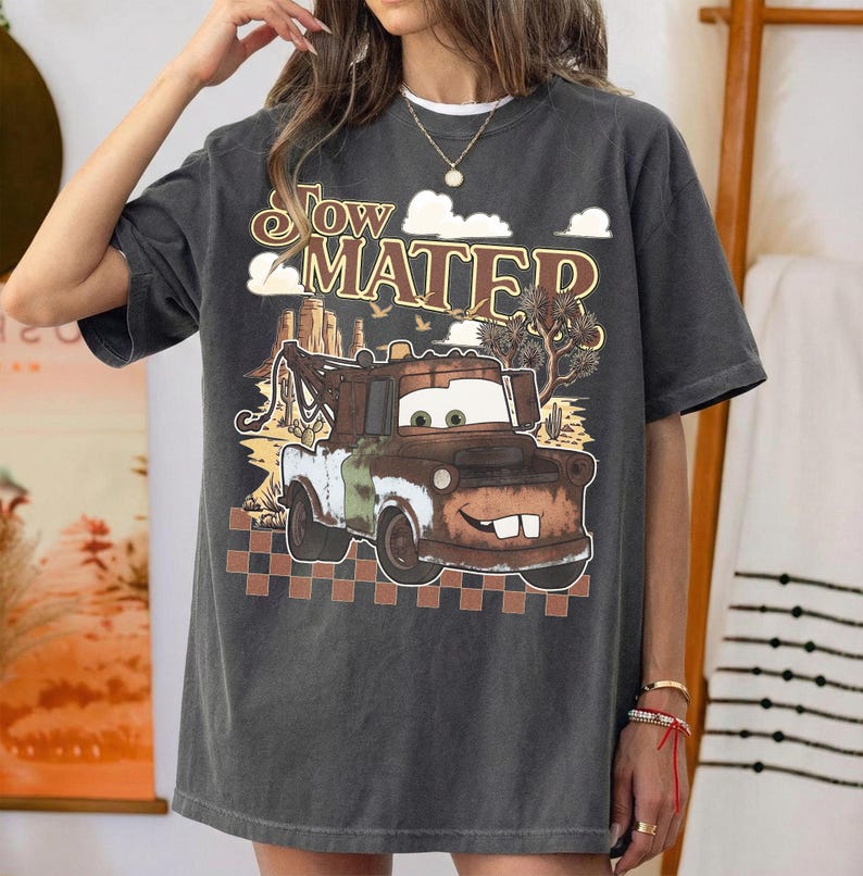 May include: A charcoal gray short-sleeve t-shirt featuring a graphic print of the character Tow Mater. The design includes the text "Tow Mater" above the truck, a desert landscape background with cacti, and a brown checkered pattern at the bottom.