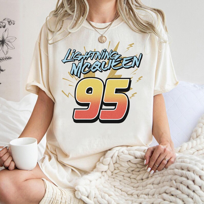 May include: A cream-coloured short-sleeve t-shirt featuring a graphic print with the text Lightning McQueen in light blue and the large number 95 in a yellow-to-red gradient. The design includes stylised lightning bolt accents in yellow.