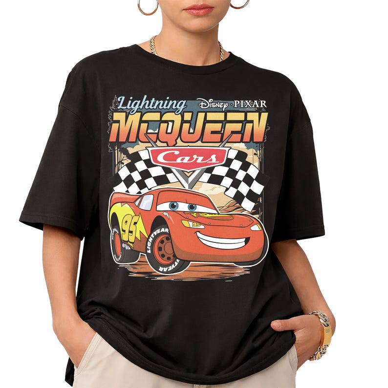 May include: A black short-sleeve t-shirt featuring a graphic print of the character Lightning McQueen from the Disney Pixar movie Cars. The design includes the text Lightning McQueen, the Cars logo, and a checkered racing flag background.