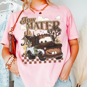 May include: A light pink t-shirt featuring a graphic of the character Tow Mater from the movie Cars. The design includes the text Tow Mater, a desert landscape with cacti, and a brown and white checkered pattern along the bottom edge.